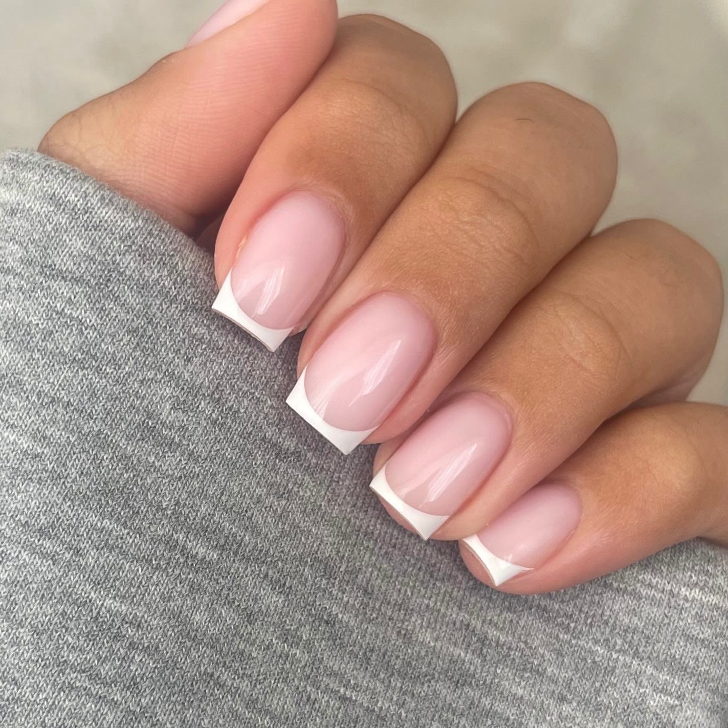Korte vierkante nagels in lichtroze met een witte French tip, hand op een grijze trui